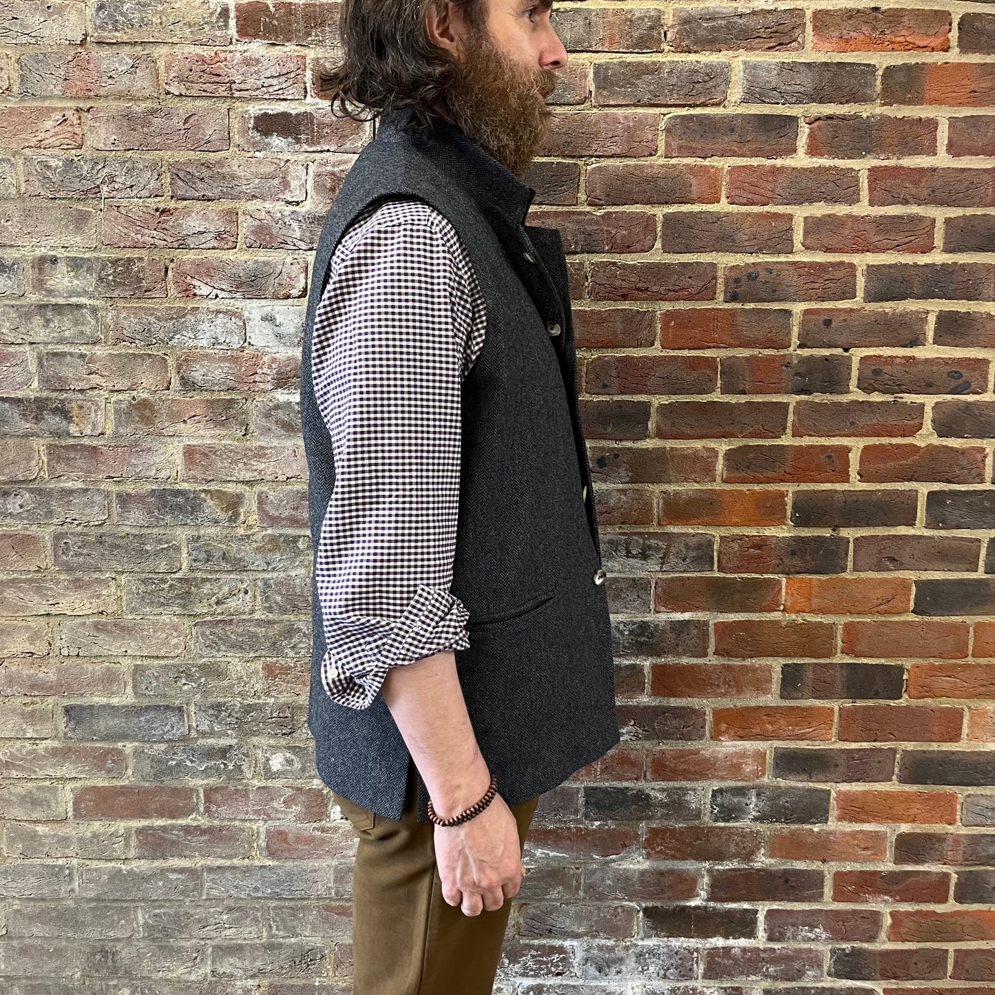 A man stood against a brick wall wearing a a grey herringbone gilet and gingham shirt