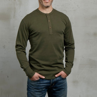 Man wearing a green long-sleeve shirt and blue jeans standing against a gray concrete wall.