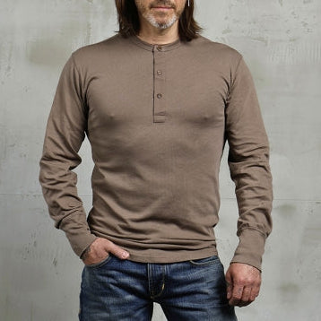 Man wearing a brown long-sleeve shirt and blue jeans against a gray concrete wall.