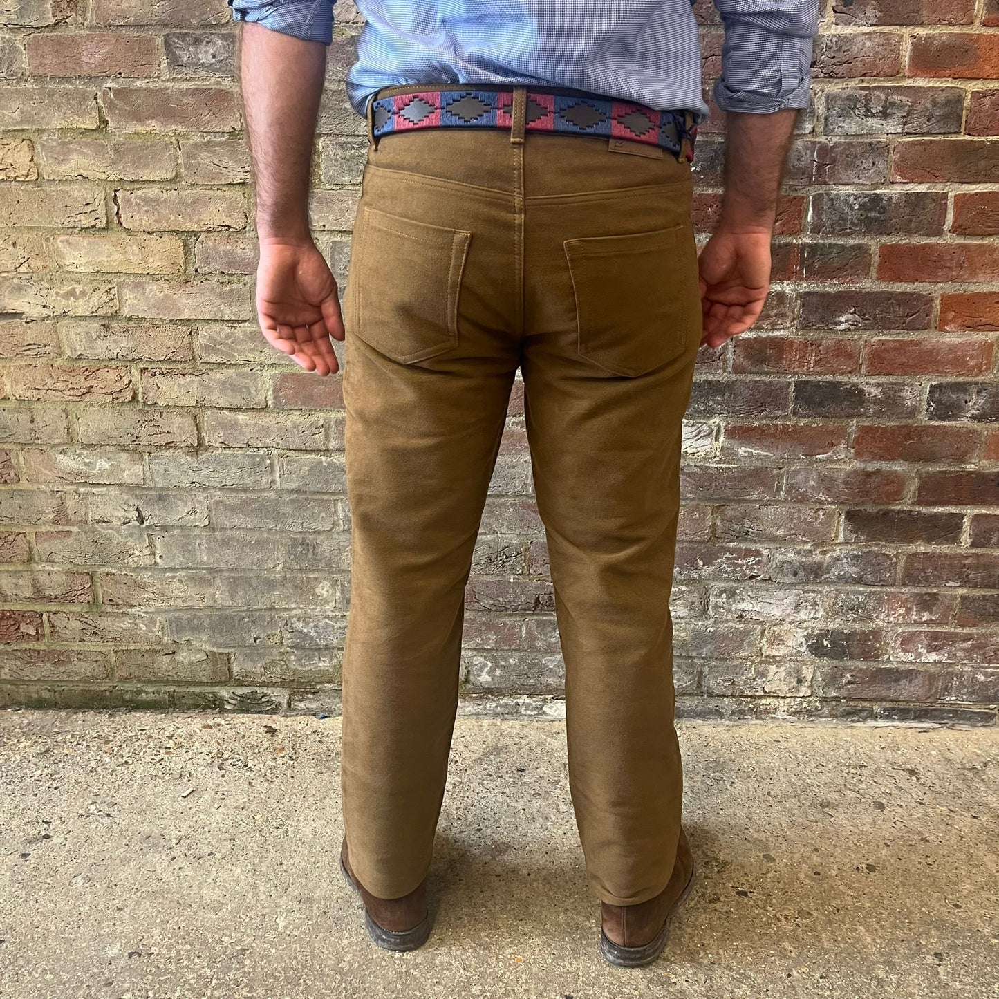 The legs of a man stood facing away from the camera against a brick wall. He is wearing a blue shirt tucked into Regent moleskins in a stone almost greenish colour with a blue and red polo belt and suede desert boots