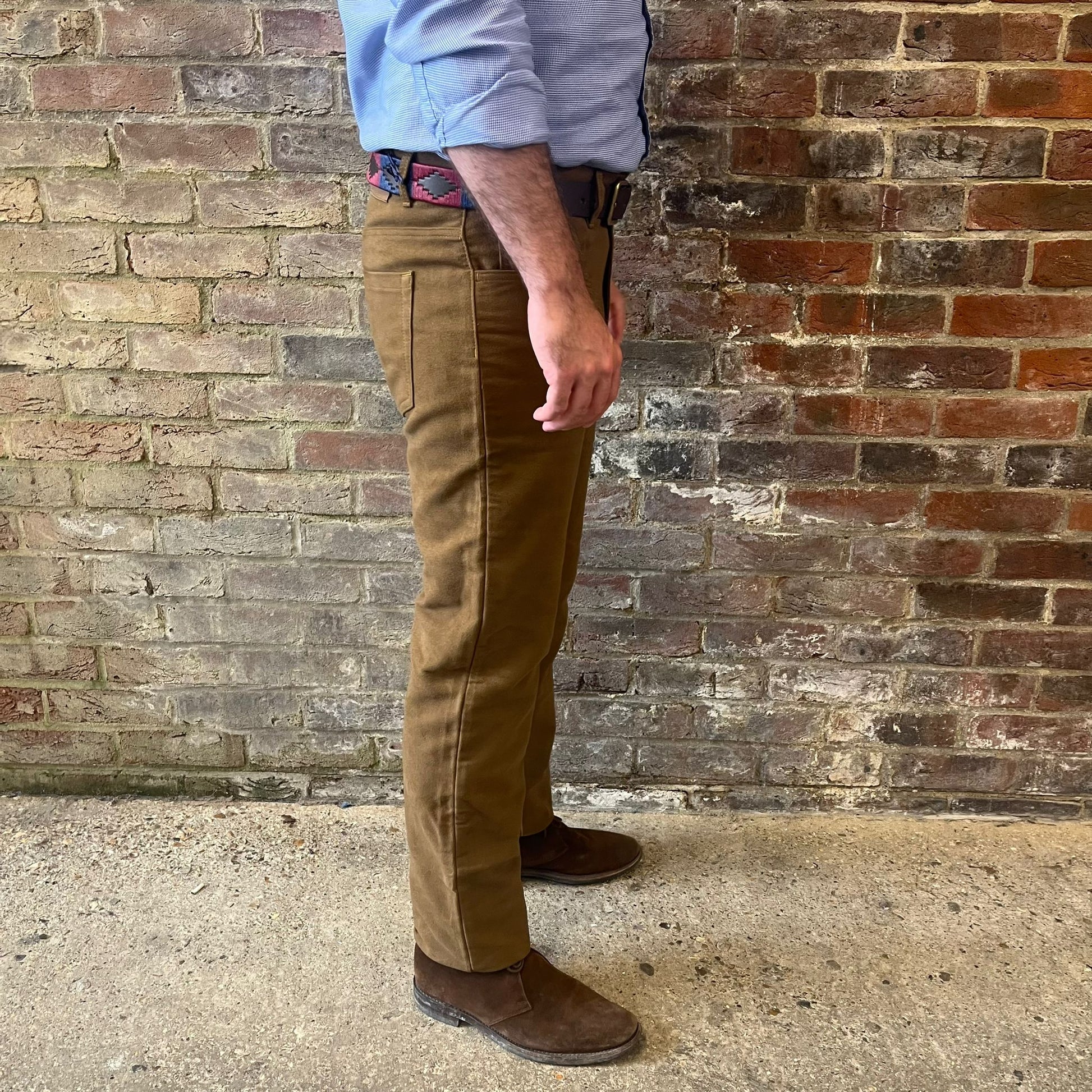 The legs of a man stood side on to the camera against a brick wall. He is wearing a blue shirt tucked into Regent moleskins in a stone almost greenish colour with a blue and red polo belt and suede desert boots