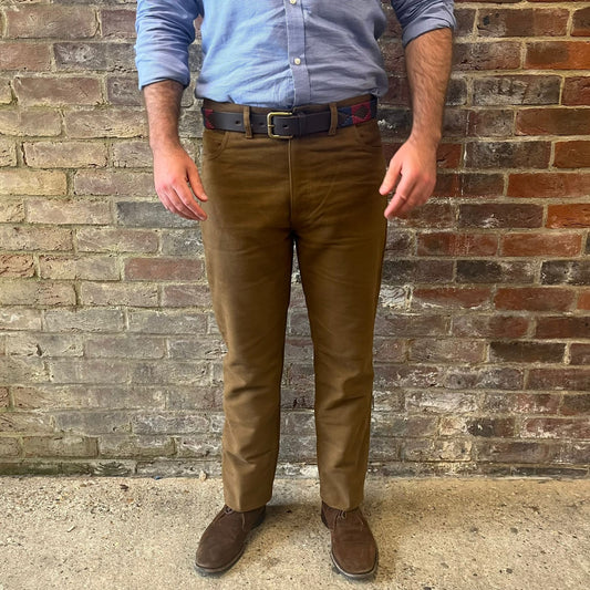 The legs of a man stood facing the camera against a brick wall. He is wearing a blue shirt tucked into Regent moleskins in a stone almost greenish colour with a blue and red polo belt and suede desert boots