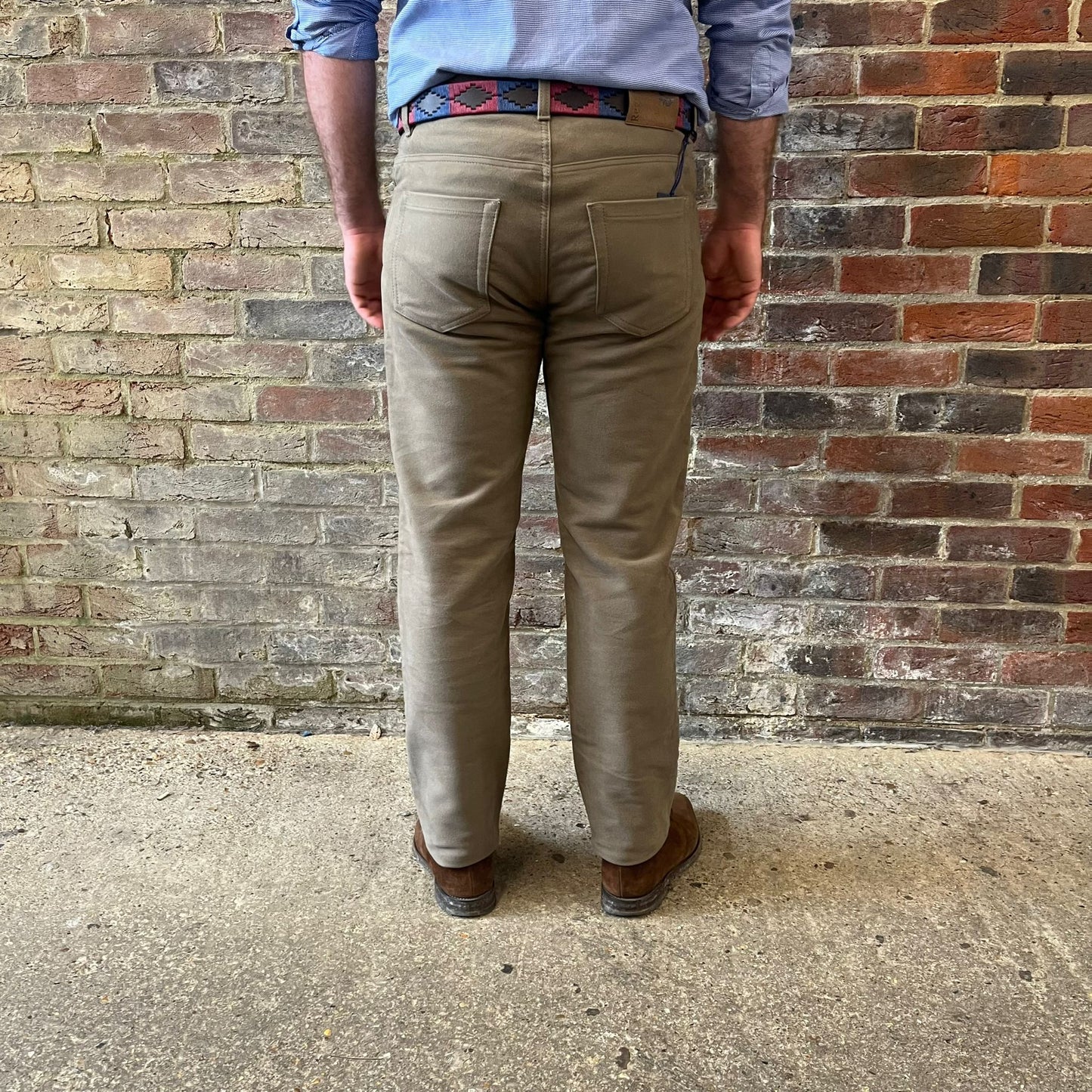 The legs of a man stood facing away from the camera against a brick wall. He is wearing a blue shirt tucked into Regent moleskins in a stone almost greenish colour with a blue and red polo belt and suede desert boots