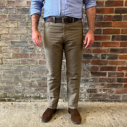 The legs of a man stood against a brick wall. He is wearing a blue shirt tucked into Regent moleskins in a stone almost greenish colour with a blue and red polo belt and suede desert boots