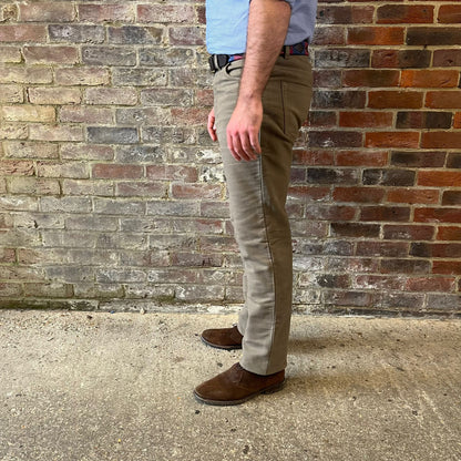 The legs of a man stood side on to the camera against a brick wall. He is wearing a blue shirt tucked into Regent moleskins in a stone almost greenish colour with a blue and red polo belt and suede desert boots