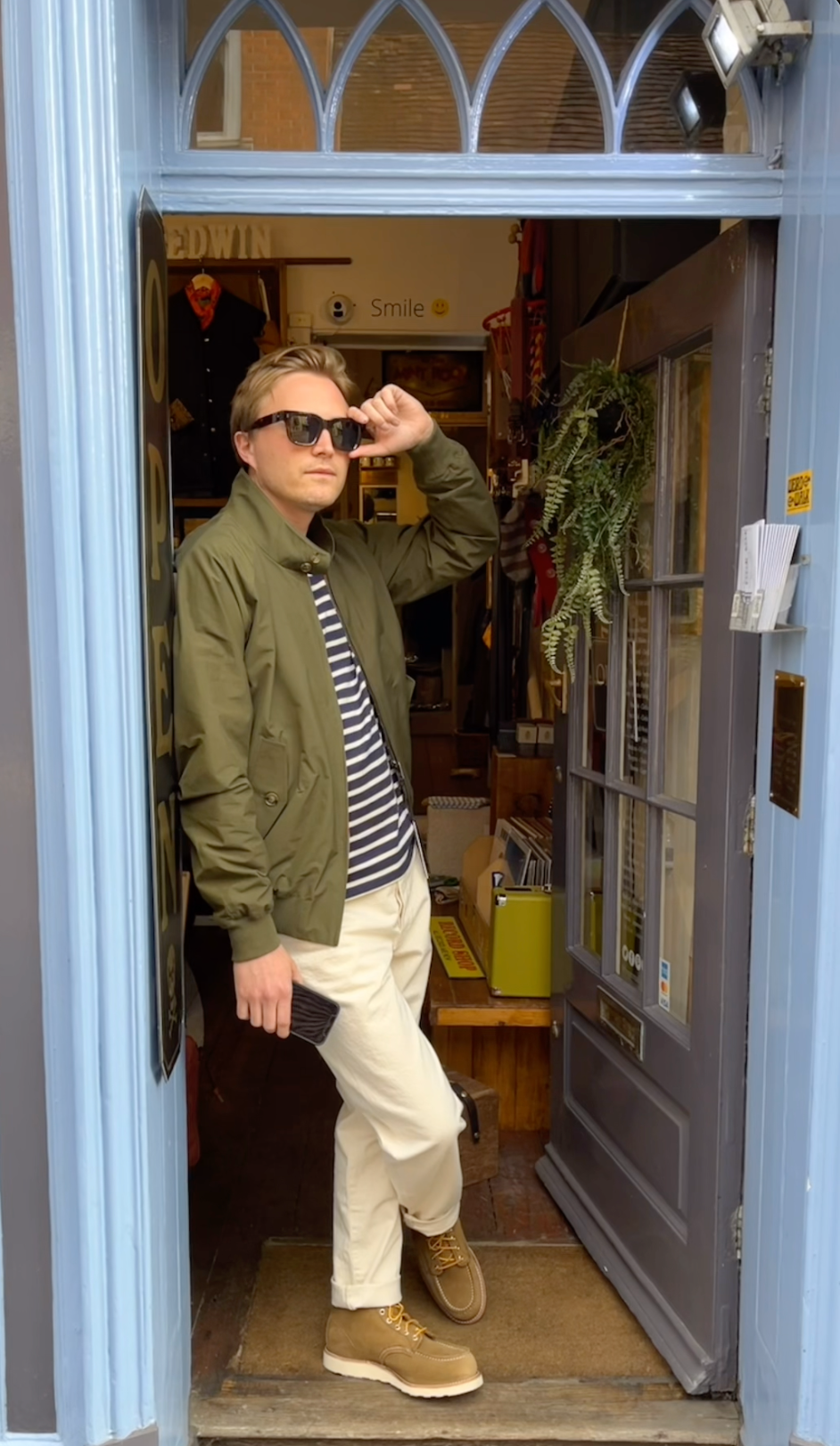 A extremely handsome man looking cool, calm and debonnaire casually standing in the doorway of a shop. He holds his Cubitts sunglasses musingly as his green baracuta harrington jacket drapes over his athletic frame pleasingly. His feet are handsomely shod in suede red wing boots and his sturdy legs adorned in charming white Edwin jeans