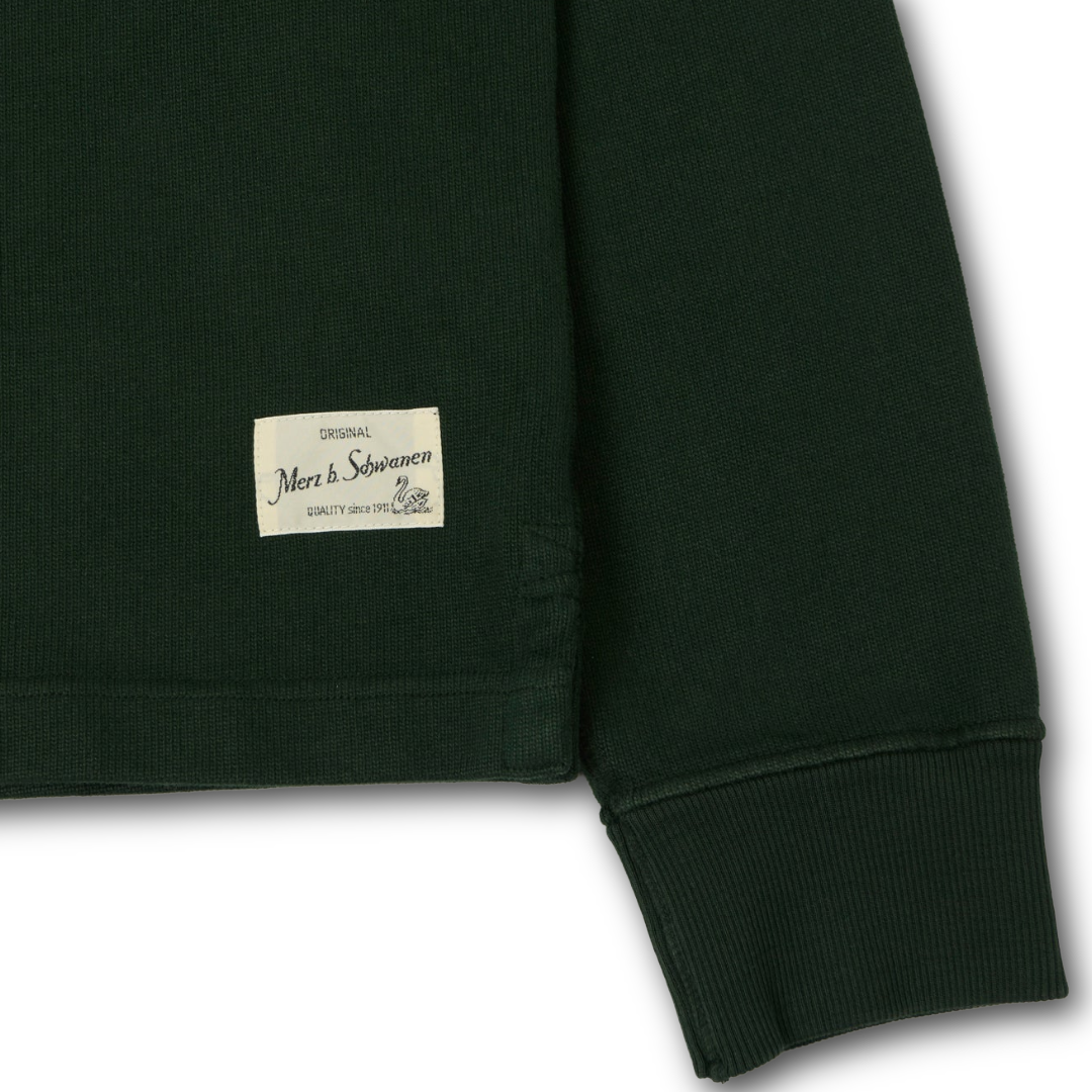 Close-up of a dark forest green ribbed knit garment, likely rugby shirt showing the sleeve cuff and a small cream sewn-in label 