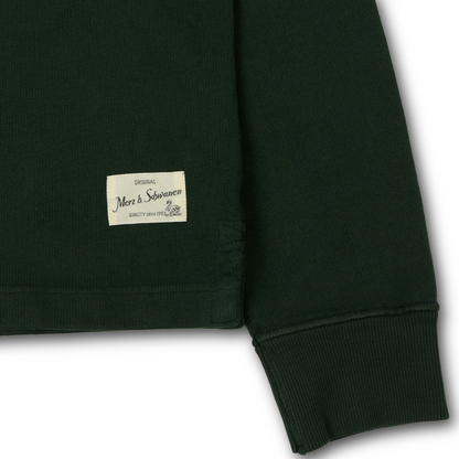 Close-up of a dark forest green ribbed knit garment, likely rugby shirt showing the sleeve cuff and a small cream sewn-in label 