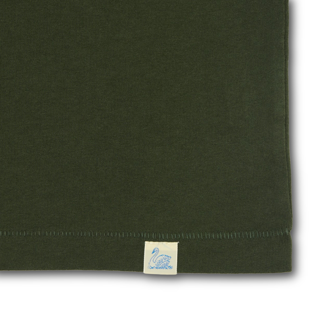 Close-up of a foresr green knit fabric product corner with a small cream fabric tag featuring a blue swan logo near the hem.