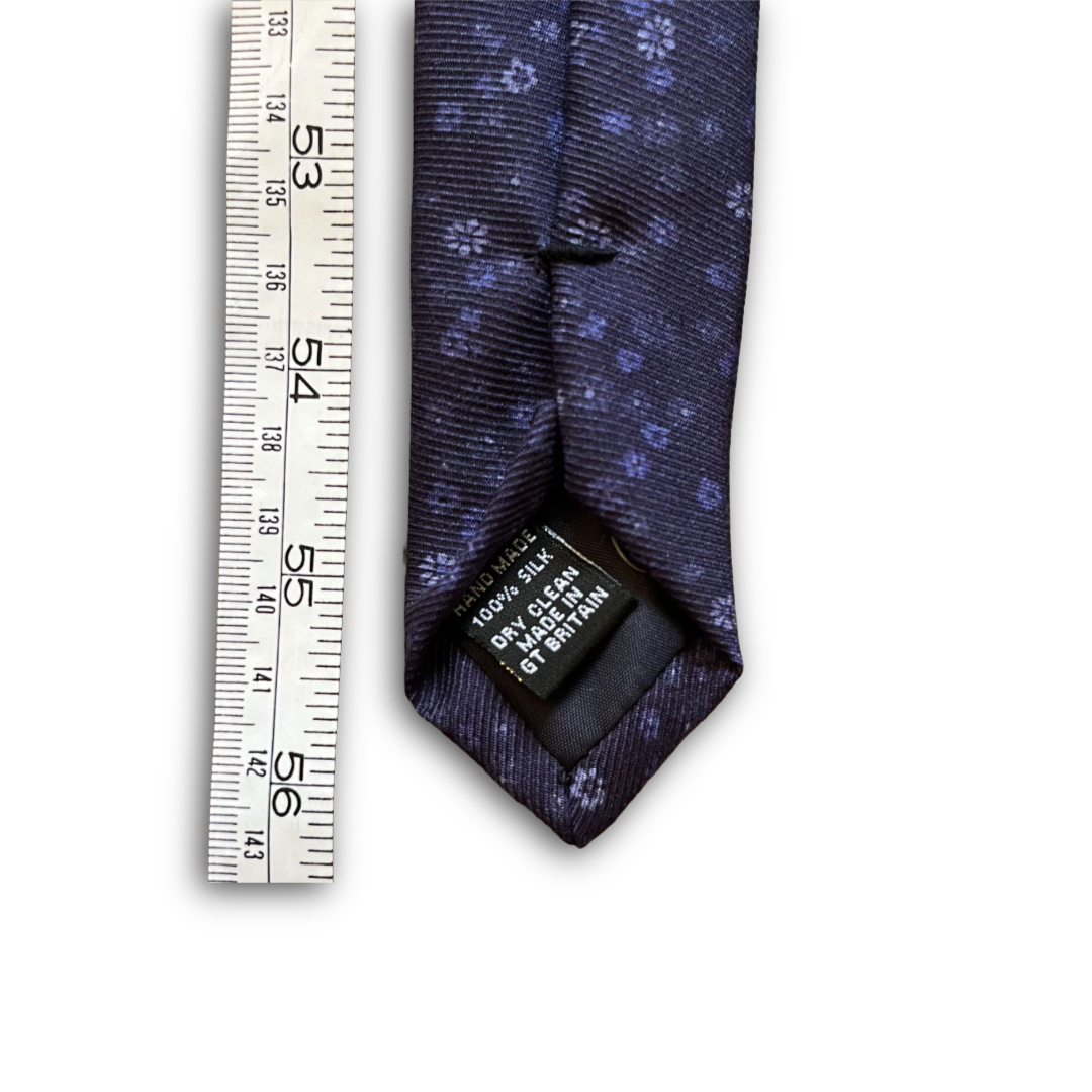 A dark navy blue necktie with a small light blue floral pattern, shown next to a white measuring ruler to indicate scale. The necktie has a black size tag visible at the collar.