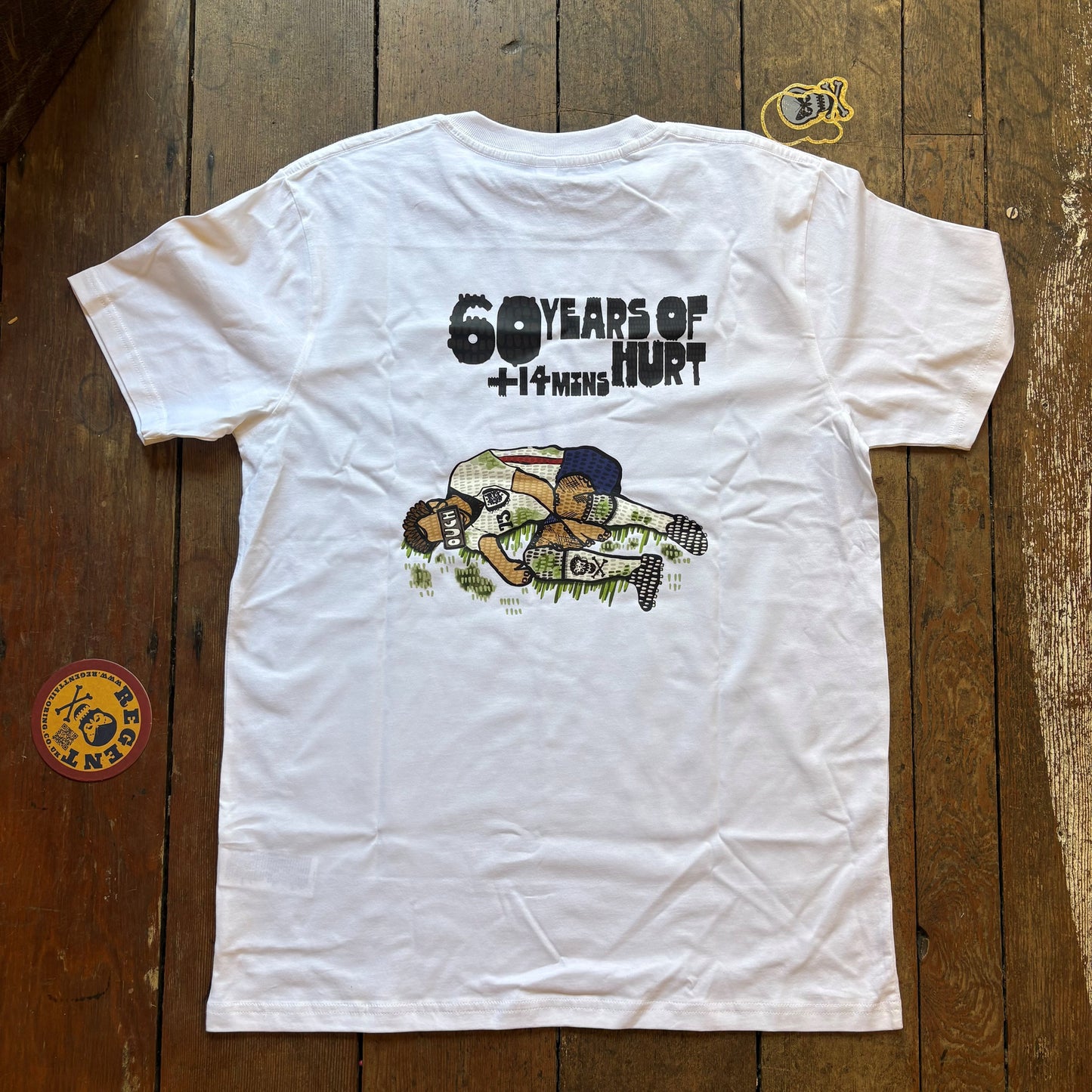 A white short-sleeve graphic T-shirt laid flat on a rustic wooden plank surface. The back of the shirt features a black text graphic reading '60 YEARS OF HURT' above a cartoon-style illustration of a football player in a white jersey and blue cap. re6gent limited edition tee shirt for the world cup 202