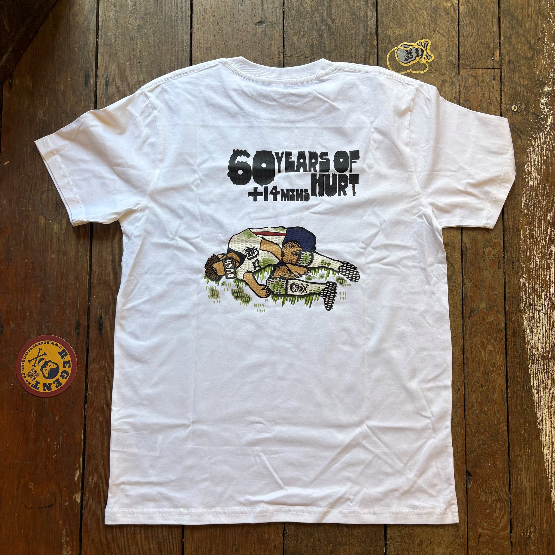 A white short-sleeve graphic T-shirt laid flat on a rustic wooden plank surface. The back of the shirt features a black text graphic reading '60 YEARS OF HURT' above a cartoon-style illustration of a football player in a white jersey and blue cap. re6gent limited edition tee shirt for the world cup 202