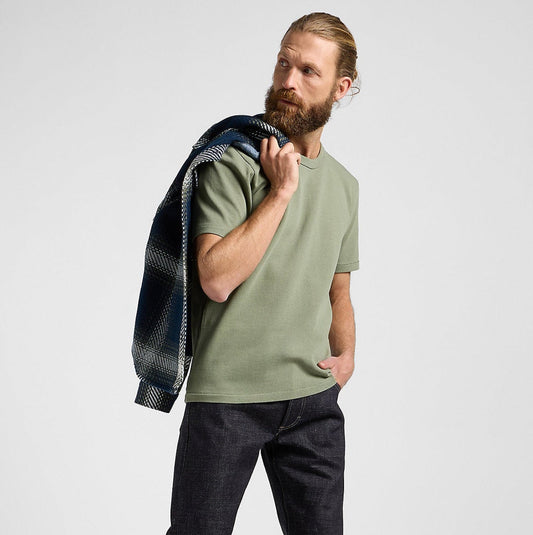 Man wearing a green t-shirt and dark jeans, holding a plaid shirt over his shoulder on a white background