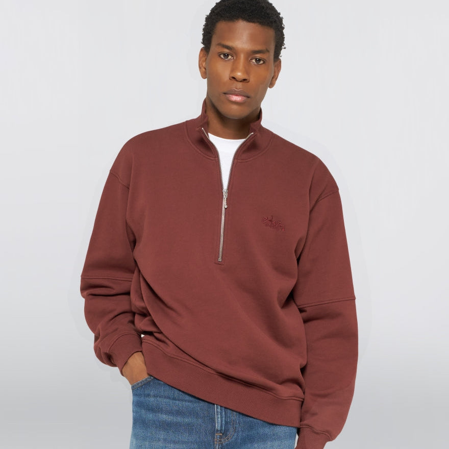 A man wearing a rust-brown quarter-zip sweatshirt with a small embroidered logo on the left chest, paired with blue jeans, against a light gray studio background.