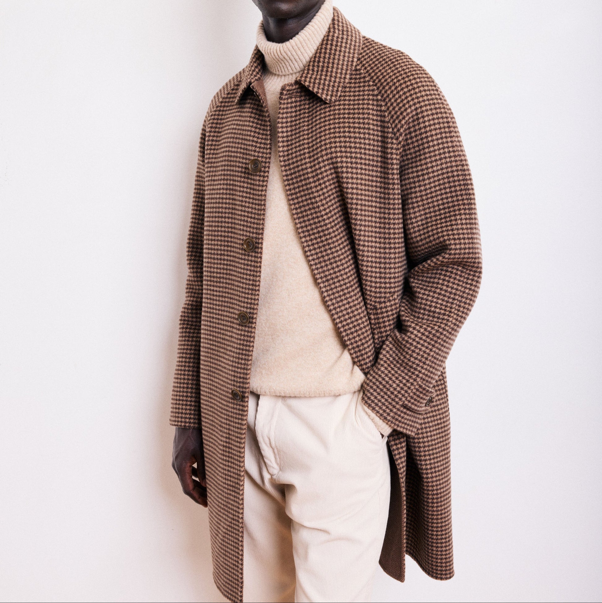 Man wearing a brown checkered coat over a beige sweater and white pants on a white background