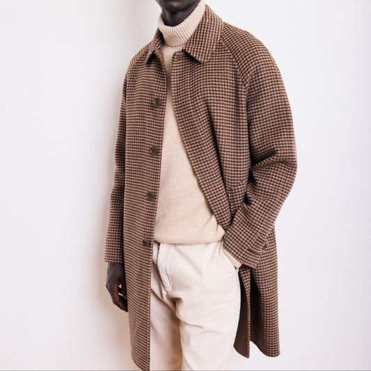 Man wearing a brown checkered coat over a beige sweater and white pants on a white background