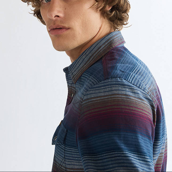 A close-up side profile of a young man with curly brown hair wearing a blue striped button-down shirt with purple and pink accents, set against a plain light gray background.