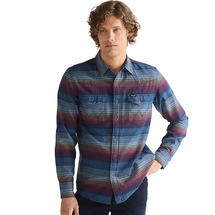 A man wearing a long-sleeve button-up shirt with a blue, red, and purple striped pattern, paired with dark jeans, photographed against a light gray studio background.