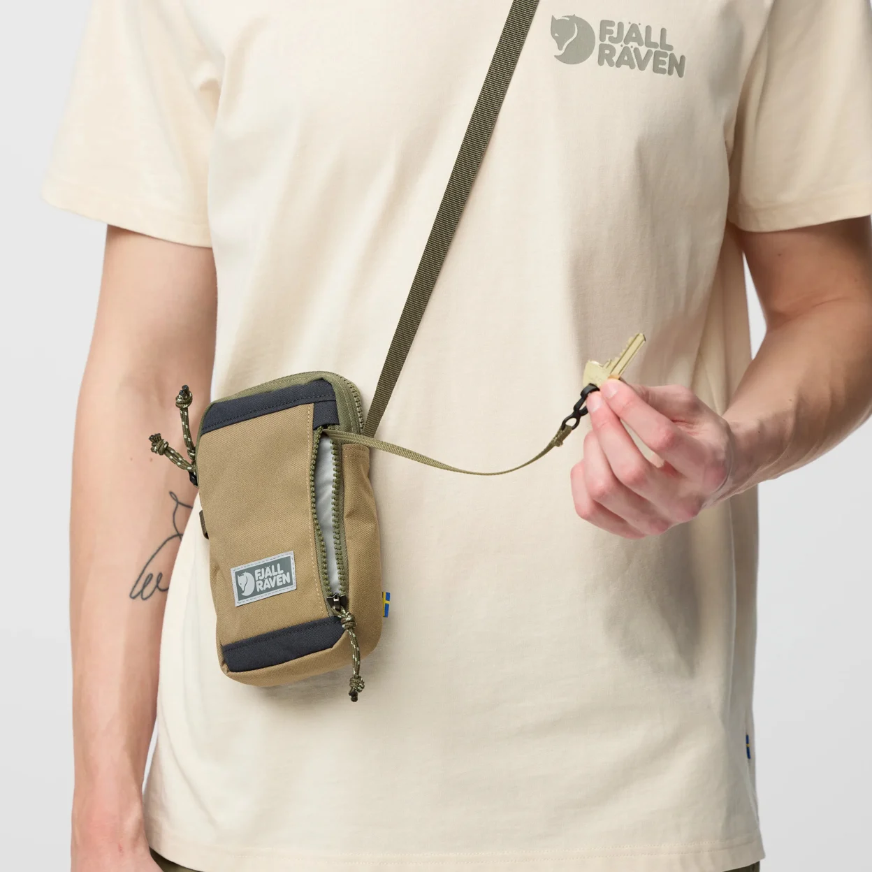Person wearing a beige t-shirt with Fjäll Räven logo, holding a matching crossbody bag.