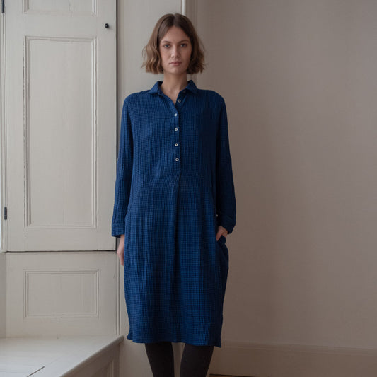 Woman wearing a blue dress standing in a room with white walls and wooden floor.