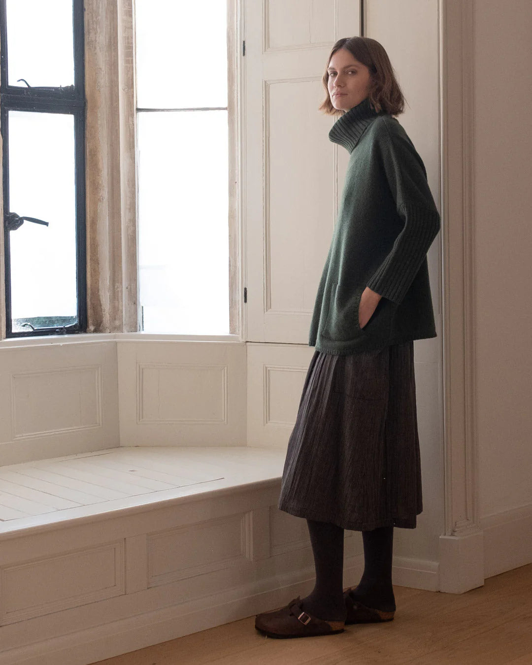 Woman in a green sweater and brown skirt standing in a room with a window seat.