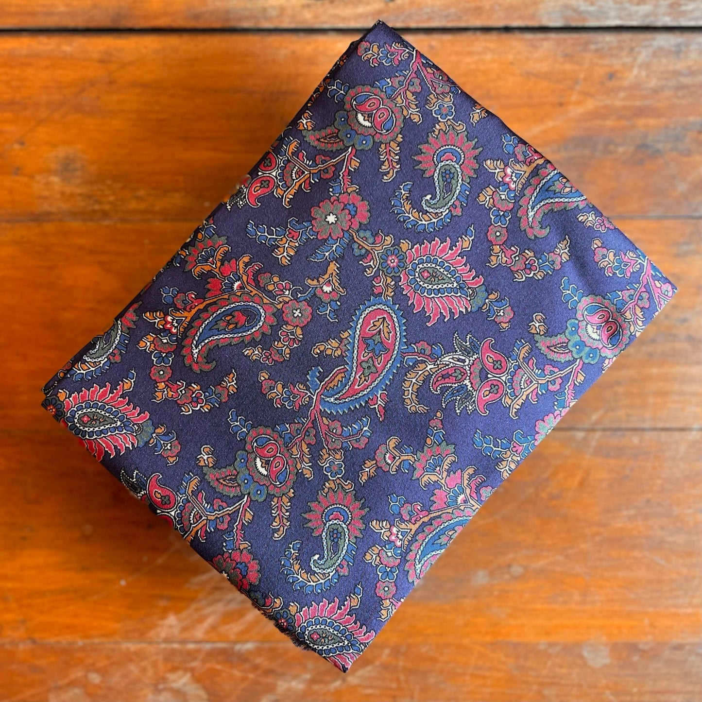 A folded blue paisley tootal scarf with red and gold accents on a wooden backdrop.